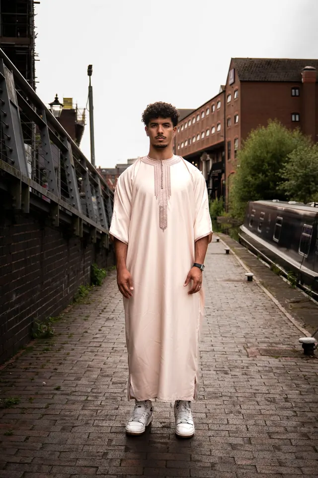 A man in an embroidered blush thobe on a canal towpath beside a narrowboat.
