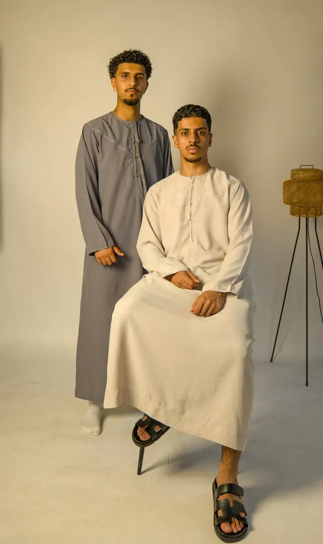 Two men in grey and stone thobes in a studio, one seated, one standing.