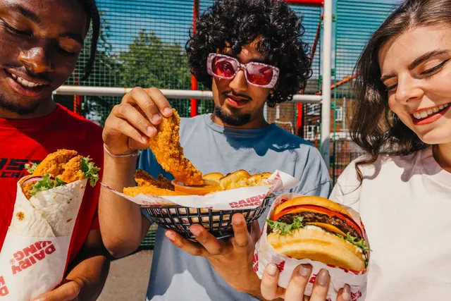 Three friends holding a wrap, a tenders basket and a burger on a sunlit court.