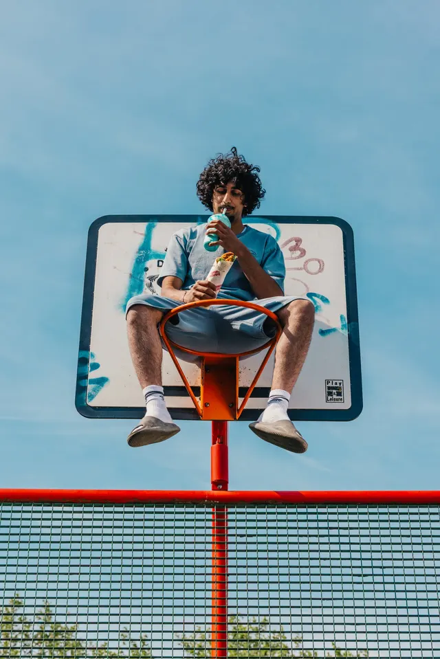 A man sitting in a basketball hoop with a wrap and a slush against a blue sky.