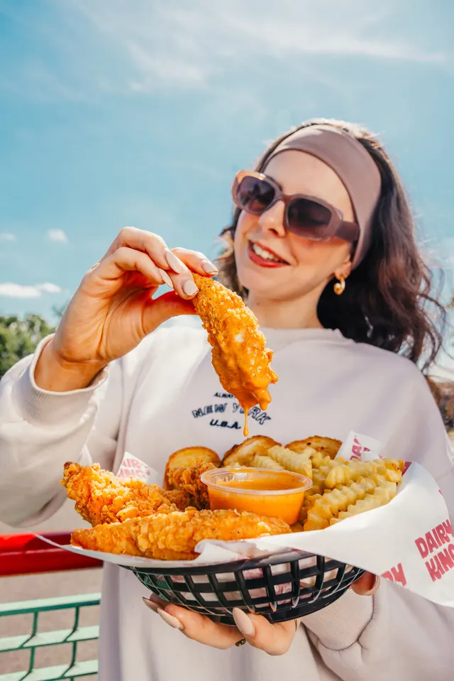 A woman lifting a chicken tender from a basket of tenders and fries.